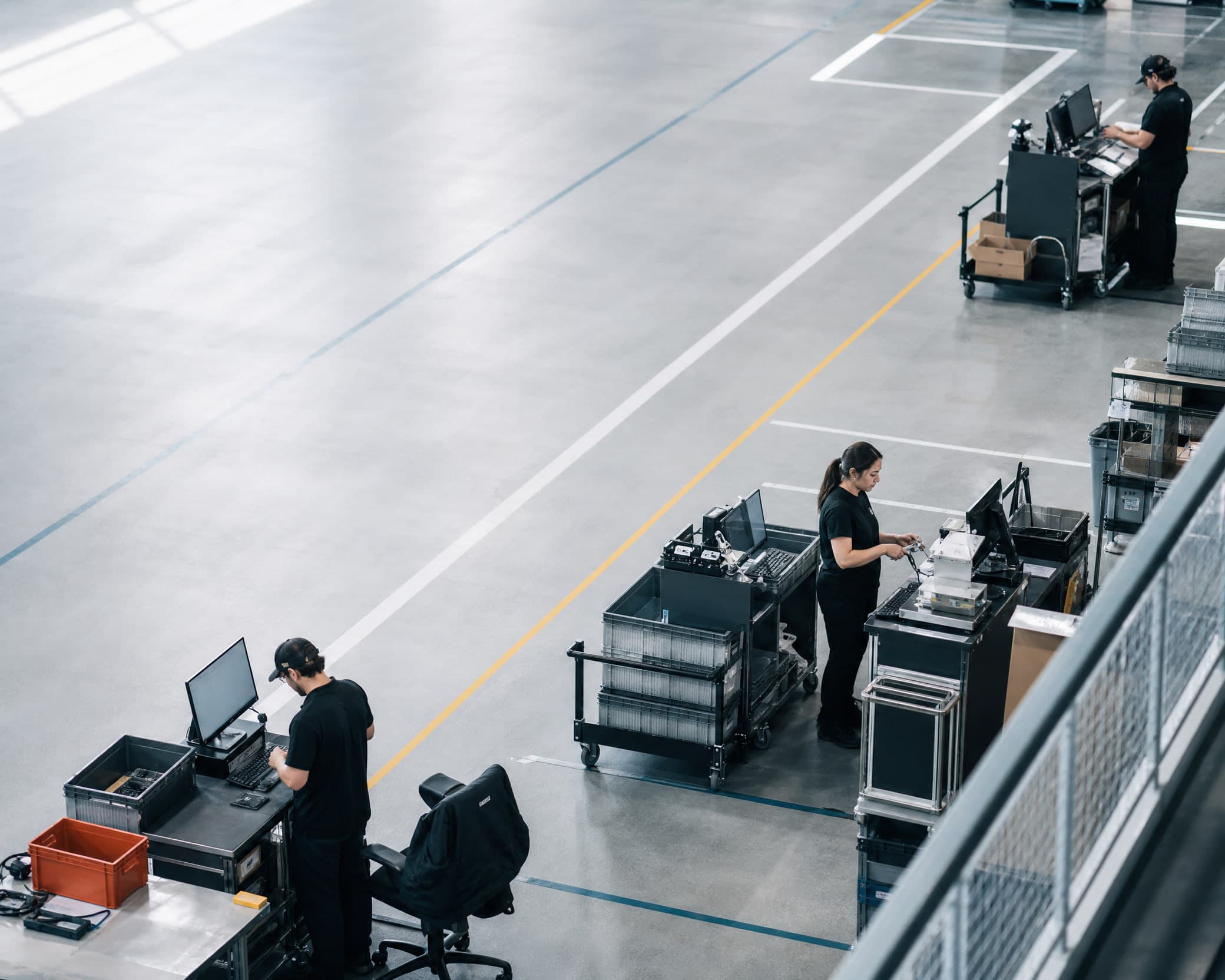Vue haute sur une plateforme logistique en pleine activité, trois personnes à des postes distincts, les lignes peintes au sol partant dans plusieurs directions.