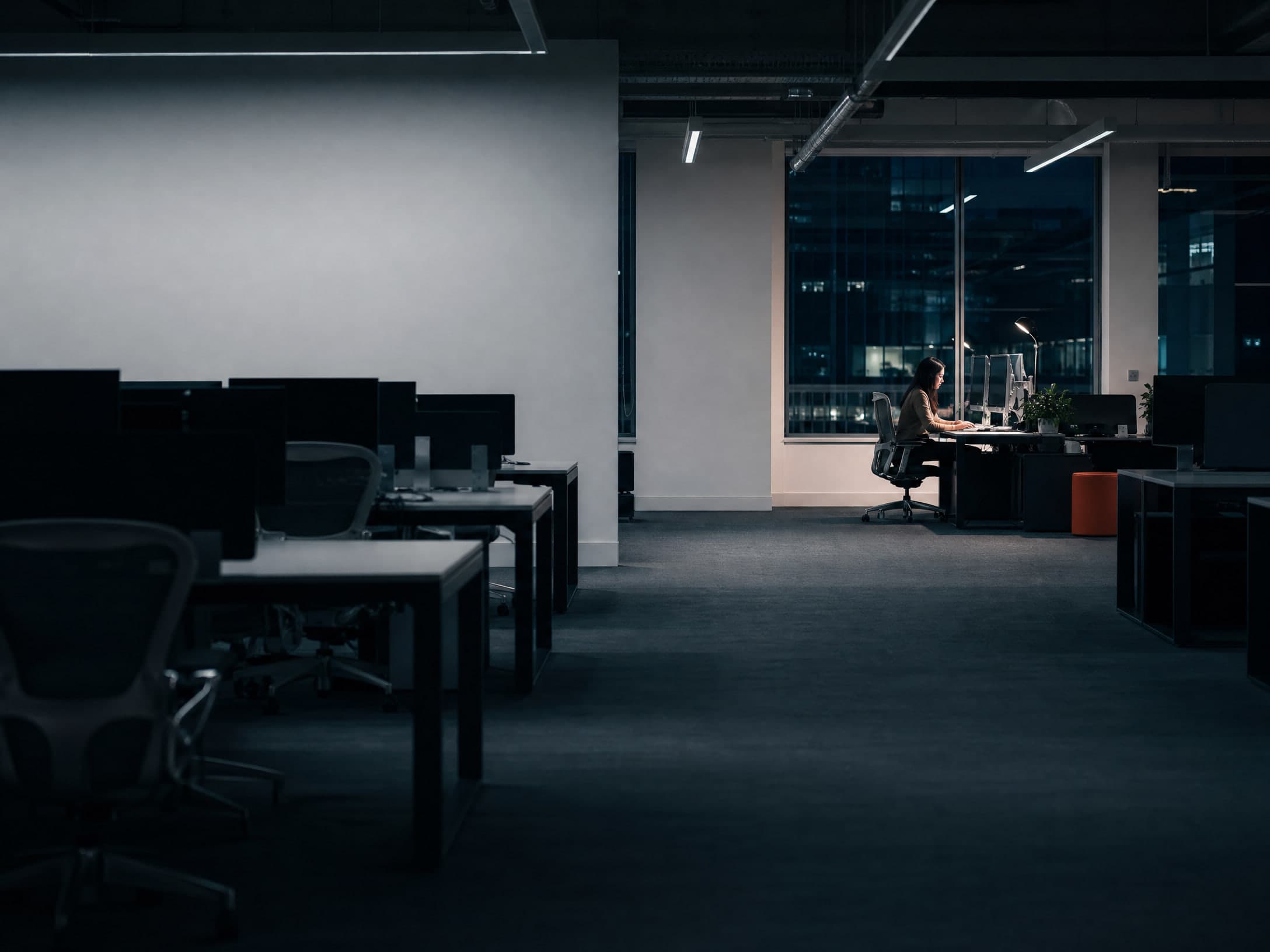 Un grand plateau de bureaux le soir, tous les postes éteints sauf un, encore occupé au fond.