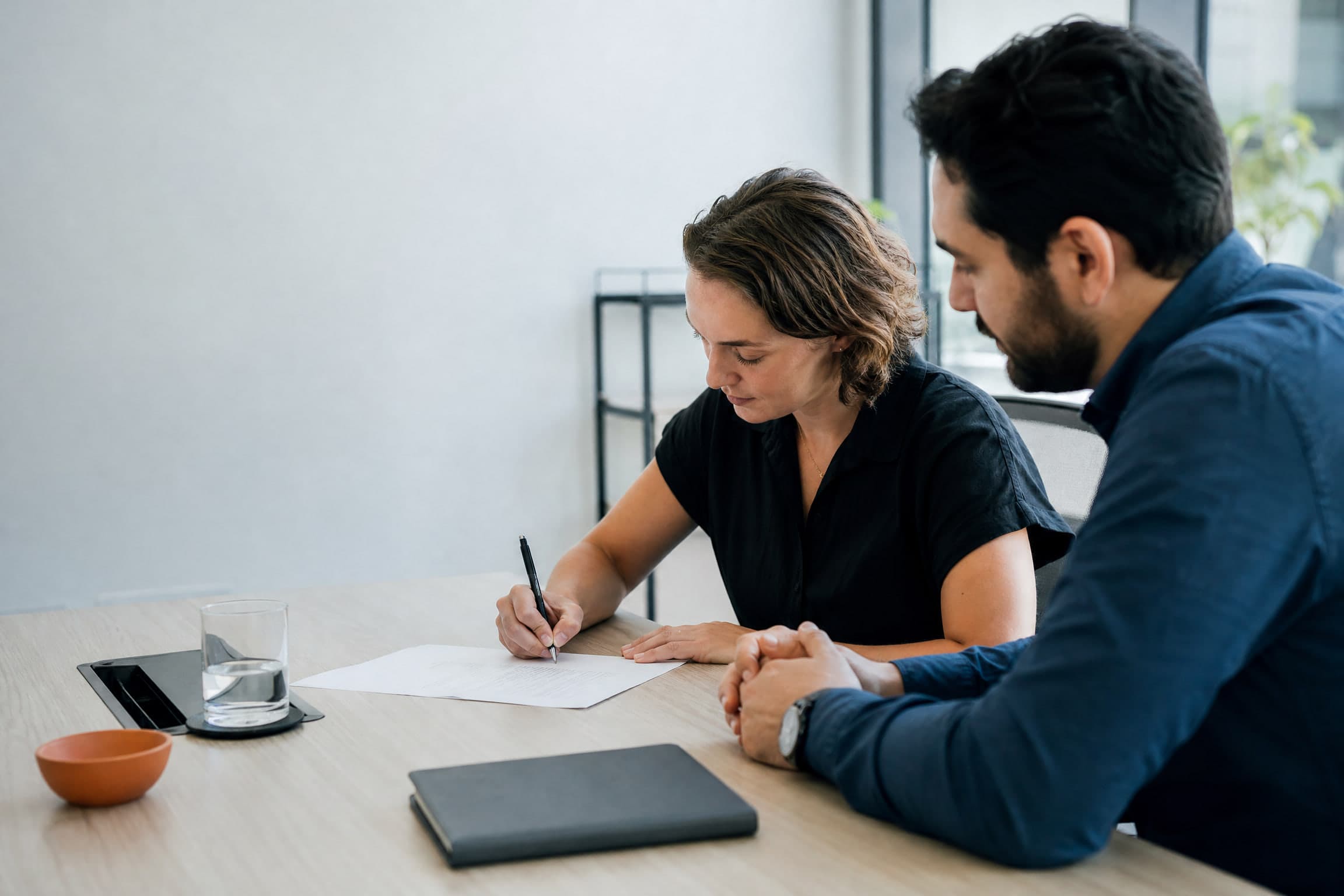 Deux personnes à un bureau, l'une signe un document imprimé pendant que l'autre regarde.